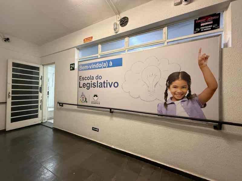 Entrada da Escola do Legislativo com um banner que diz 'Bem-vindo(a) à Escola do Legislativo'. À direita, uma menina sorridente levanta a mão, simbolizando aprendizado e participação.
