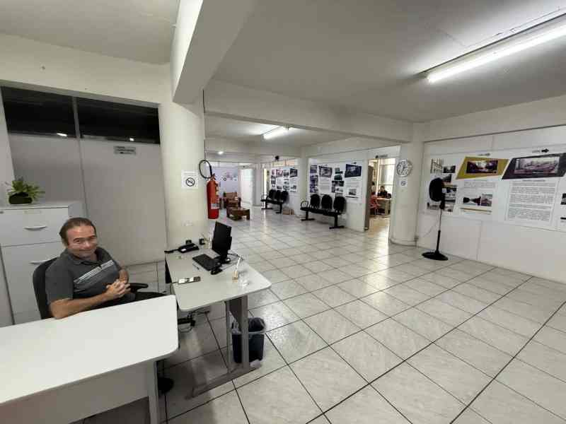 Interior da Escola do Legislativo com um funcionário sentado em uma mesa ao fundo. O espaço é aberto, com composta por computadores, cadeiras e painéis informativos nas paredes.