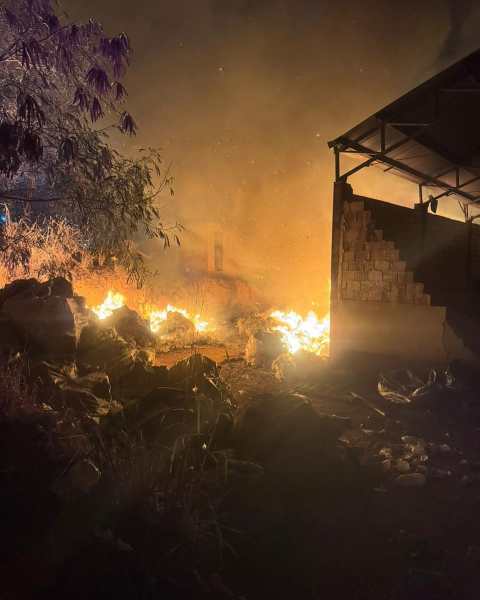 Incêndio em área de disposição de resíduos sólidos, com chamas visíveis e fumaça densa ao fundo, perto de um galpão parcialmente danificado.