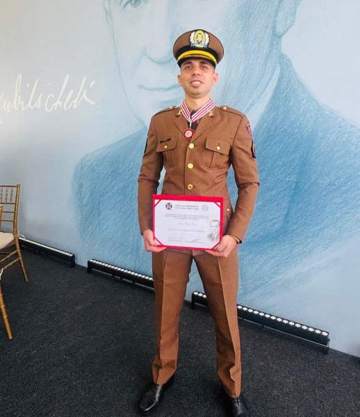 Cabo Lucas Costa Neto, em uniforme da Polícia Militar, segurando um certificado de condecoração com a Medalha Presidente Juscelino Kubitschek em uma cerimônia na presença de um retrato ao fundo.