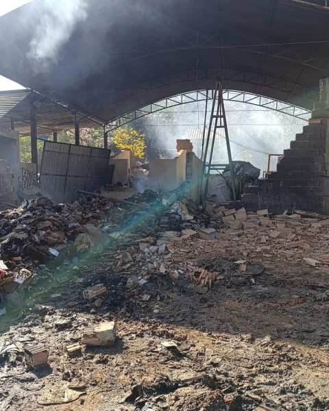 Área atingida por incêndio em um galpão com resíduos sólidos, fumaça visível e escombros ao redor.