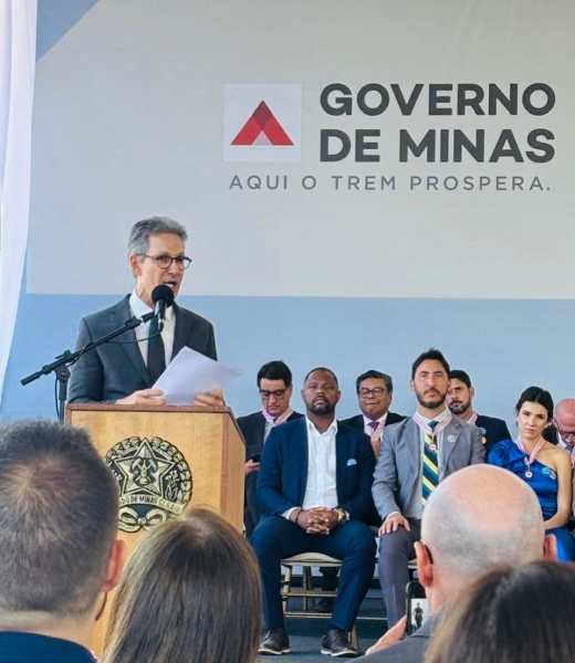 Um orador discursa em uma cerimônia do Governo de Minas, com uma plateia atenta ao fundo, incluindo autoridades e convidadas, em um ambiente institucional.