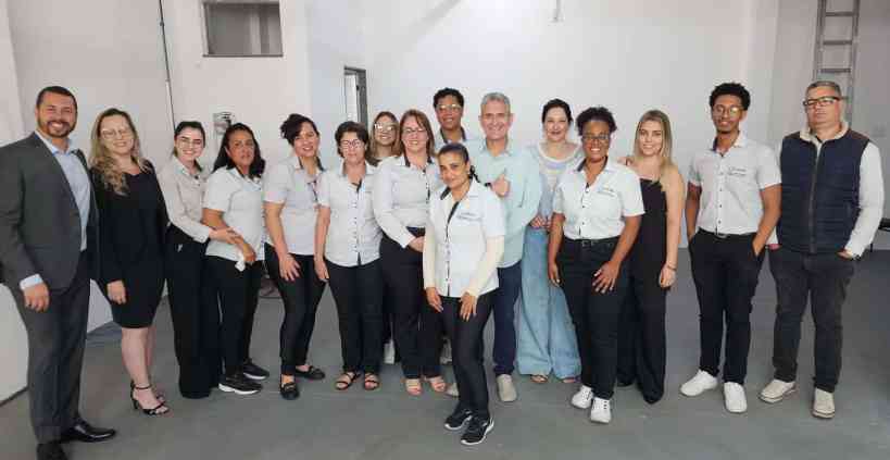 Grupo de pessoas posando para foto em um evento, vestidos com camisetas ou uniformes padronizados, em um ambiente interno com paredes claras.