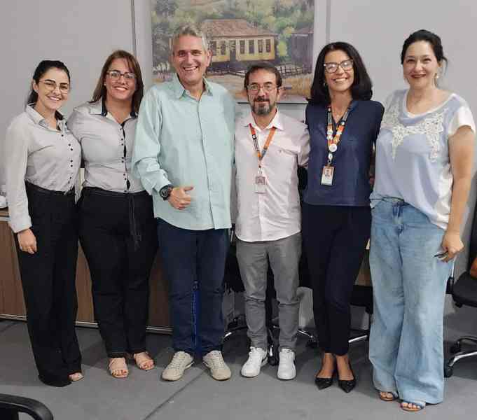 Foto de um grupo de seis pessoas em um ambiente interno, posando para a câmera. As pessoas estão sorrindo e a decoração inclui uma pintura ao fundo. Todos estão vestindo roupas casuais e formais.