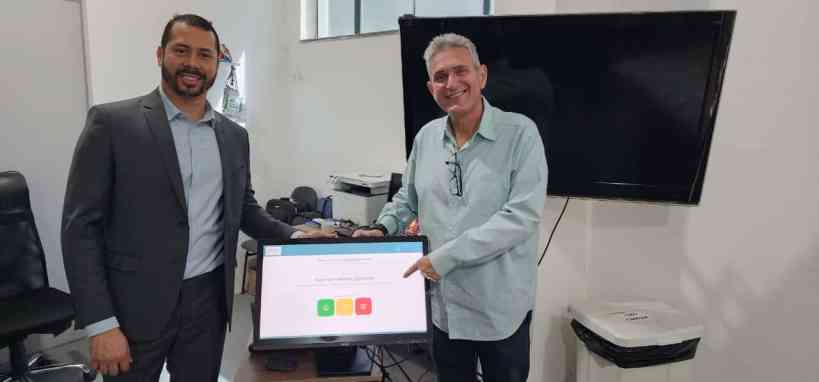 Dois homens sorridentes em um ambiente de escritório, um deles segurando uma tela de computador que exibe um sistema de autoatendimento para agendamento de serviços de saúde.