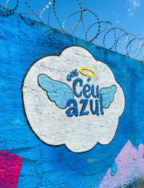 Mural colorido com o nome da ONG Céu Azul, rodeado por uma nuvem branca e com asas, em um fundo azul. O design reflete uma atmosfera acolhedora e positiva.