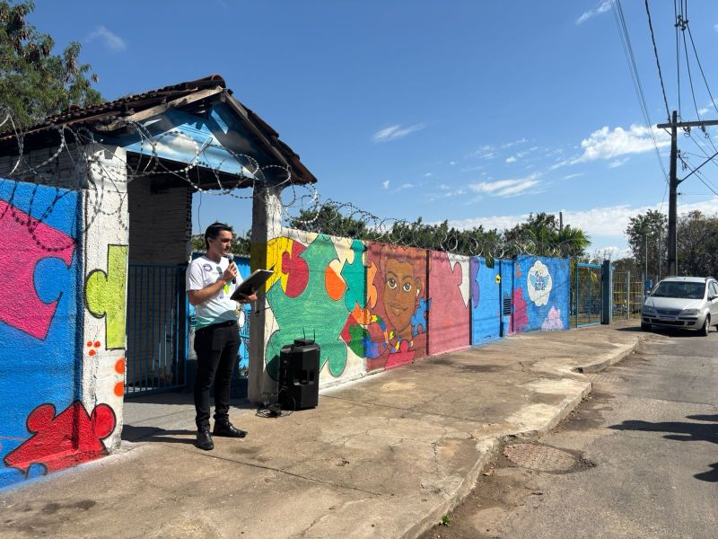 Um homem em pé, lendo um documento, em frente a uma parede pintada com diversos quebra-cabeças coloridos e um mural de uma criança. É um dia ensolarado, com algumas nuvens no céu.