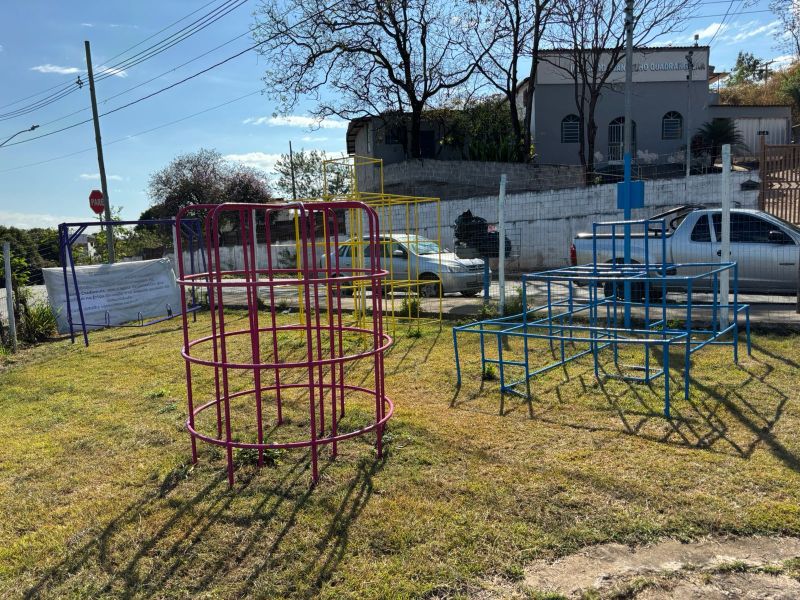 Espaço revitalizado com estruturas coloridas de playground, como arcos e balanços, cercado por gramado e uma parede ao fundo, em um dia ensolarado.