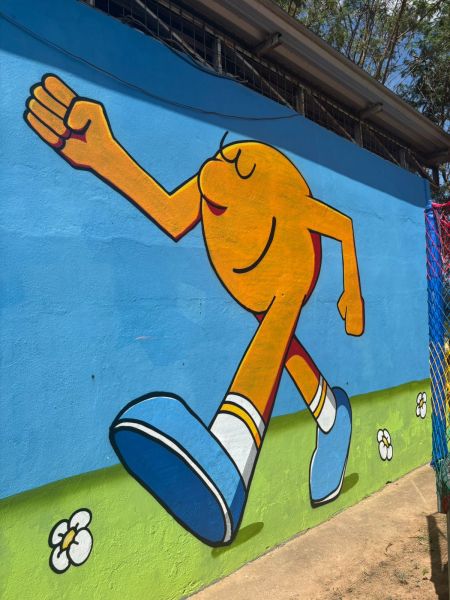 Pintura mural colorida de uma figura sorridente caminhando em um fundo azul, com flores brancas na grama verde embaixo.