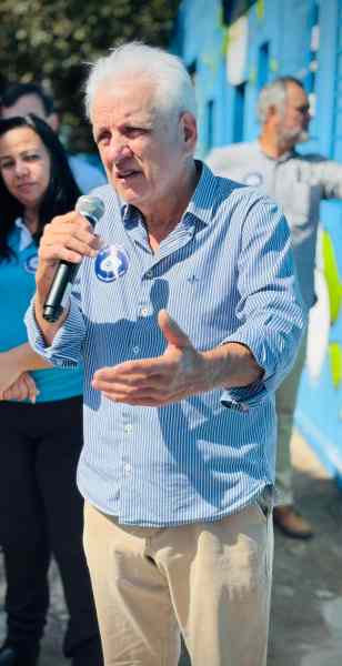 Homem com cabelo grisalho e camiseta listrada azul falando ao microfone em uma cerimônia de revitalização, com um fundo de paredes azuis.