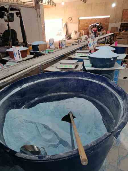 Interior de uma fábrica clandestina de sabão em pó falsificado, mostrando baldes com pó e caixas de produtos embalados.