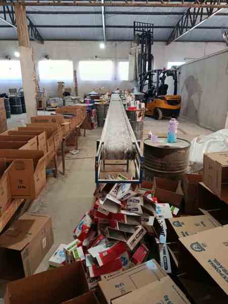 Interior de uma fábrica clandestina de sabão em pó falsificado, com caixas empilhadas e equipamentos de produção visíveis.