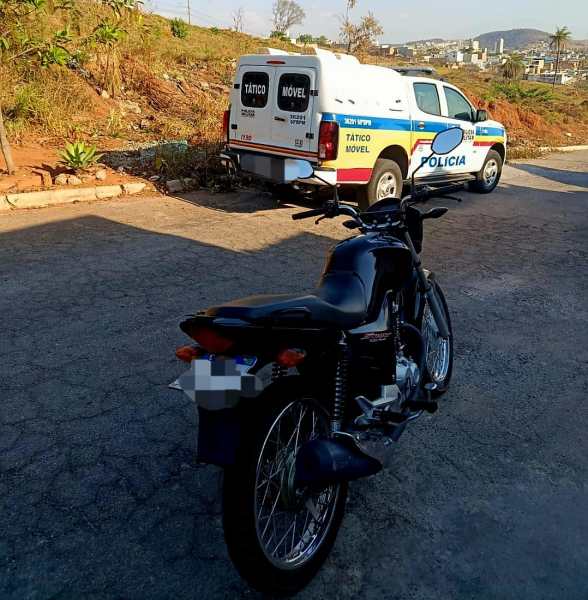Imagem de uma motocicleta preta estacionada com um veículo da Polícia Militar ao fundo, em uma área urbana. A cena reflete a fiscalização de trânsito durante a Operação Cavalo de Aço.