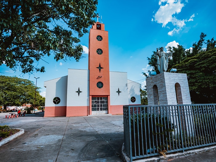 Igreja situada no bairro Interlagos, em Divinópolis, com estrutura branca e detalhes em laranja, cercada por árvores e um céu azul.