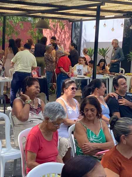 Grupo de pessoas sentadas em um evento ao ar livre, com mesas e decoração simples, desfrutando de uma atividade cultural.