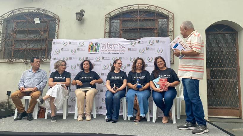 Grupo de pessoas sentadas em um palco durante o evento Fliarte, com um banner ao fundo, promovendo a interação cultural e literária.