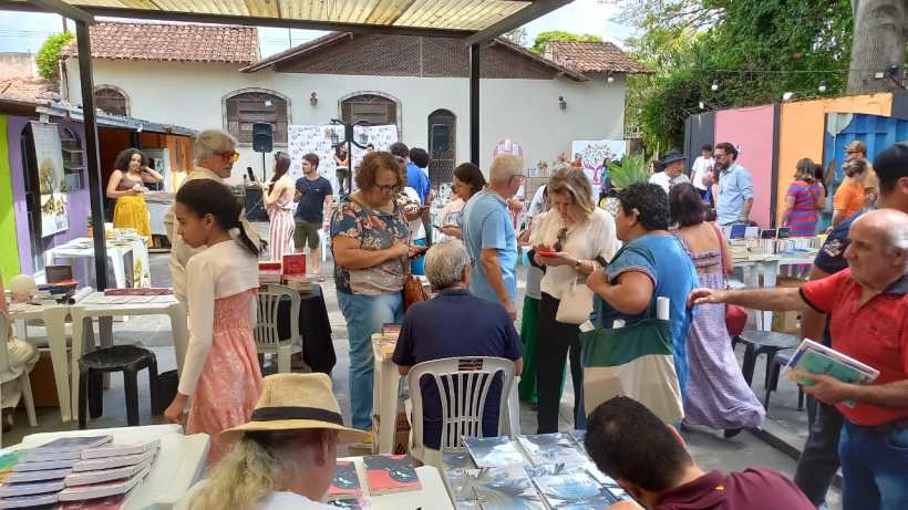 Evento cultural com diversas pessoas interagindo em um espaço ao ar livre, com mesas cheias de livros e materiais literários. Ao fundo, há apresentações e um ambiente animado.