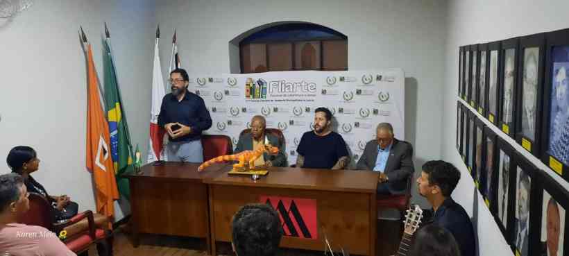 Mesa de apresentação do Fliarte com oradores e participantes, bandeiras ao fundo e fotos expostas na parede.