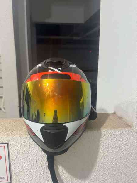 Um capacete de motociclismo com vis visor espelhado, apoiado em uma superfície branca. Ao fundo, é possível ver uma parede e uma luz refletida.