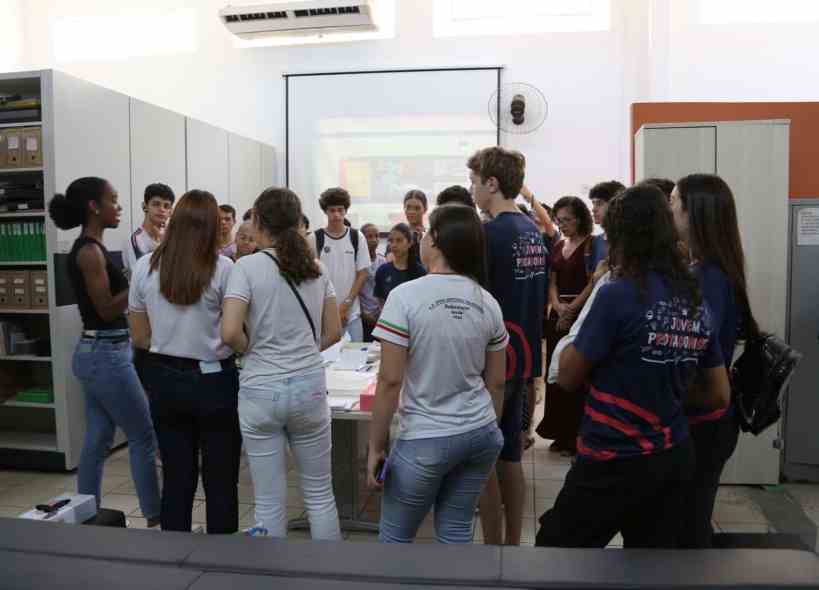 Estudantes participando de uma apresentação em grupo na Feira de Profissões da UEMG, com um palestrante à frente e uma tela ao fundo.