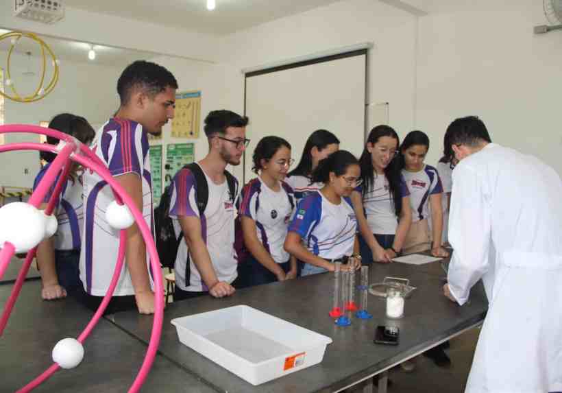 Estudantes participando de uma atividade prática em laboratório, observando atentamente um instrutor que demonstra um experimento utilizando materiais de ciências.