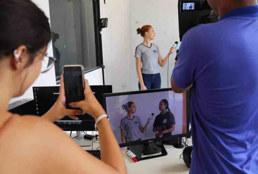 Estudantes participando de uma gravação em um estúdio, com uma pessoa sendo filmada enquanto outra a observa com um celular na mão.