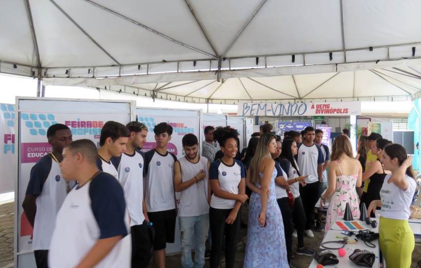 Um grupo de estudantes participando da Feira de Profissões da UEMG Divinópolis, com estandes informativos ao fundo.