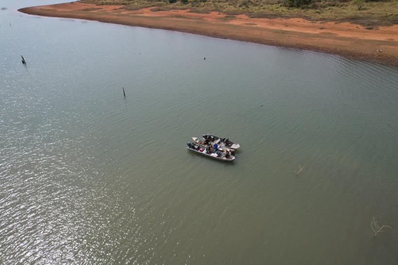 Uma vista aérea de uma embarcação com várias pessoas navegando em um lago, cercado por uma costa de terra avermelhada.