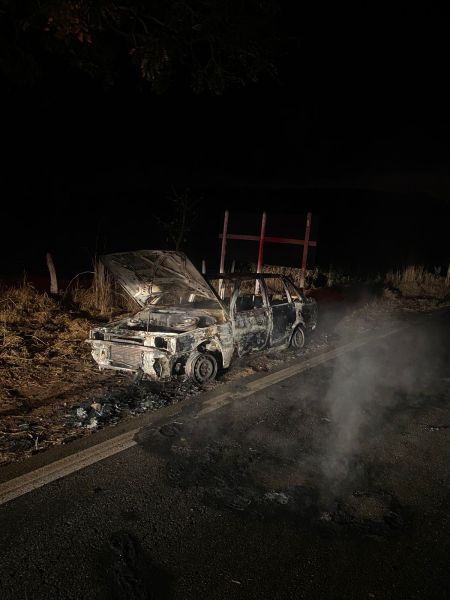 Veículo queimado à beira da estrada, com fumaça saindo do motor, durante a noite.
