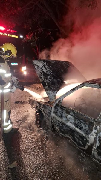 Bombeiro combatendo incêndio em veículo com fumaça e chamas na BR-332 à noite.