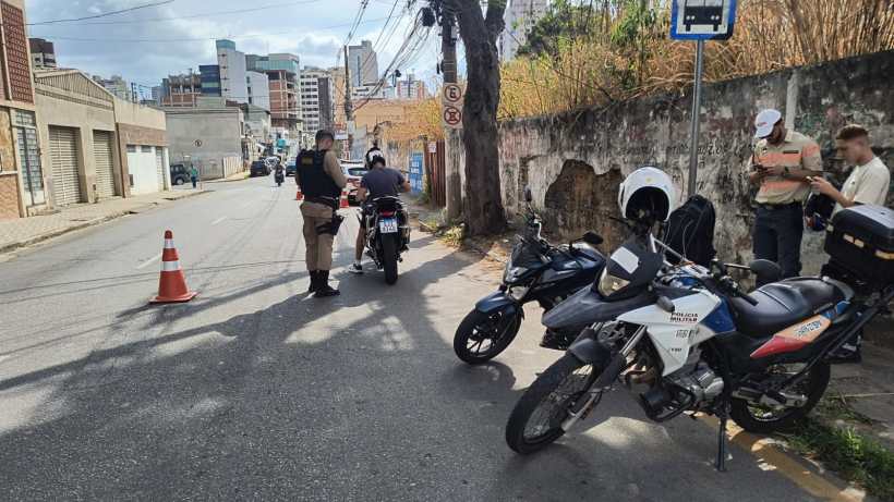 Polícia Militar e Secretaria Municipal de Trânsito realizando blitz em Divinópolis, com agentes abordando motociclistas e verificando documentos, enquanto veículos estão estacionados ao lado da rua.