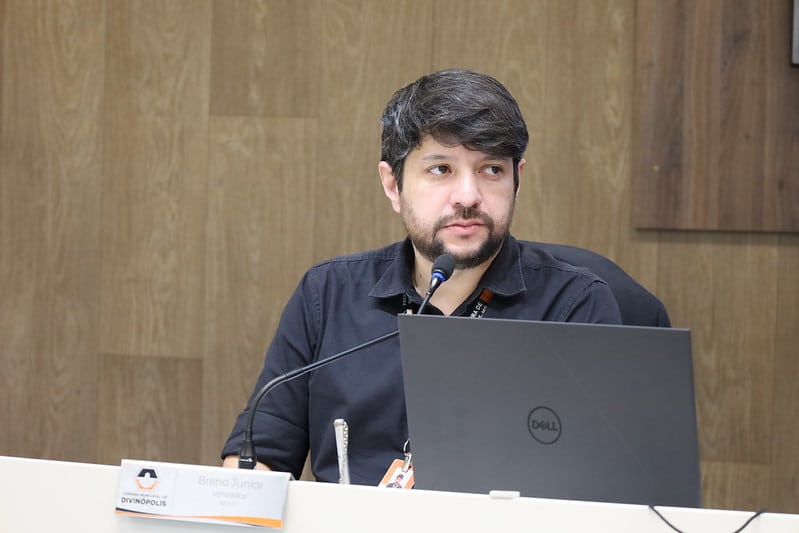 Homem com cabelo escuro e barba, sentado em mesa de câmara municipal, com laptop à sua frente, participando de audiência pública.