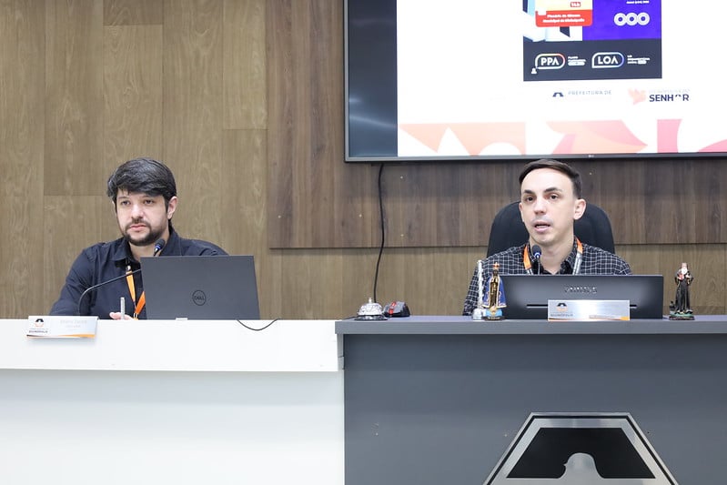 Dois homens em uma mesa de apresentação, um deles com um laptop e um microfone, explicando os detalhes do orçamento municipal para 2026.