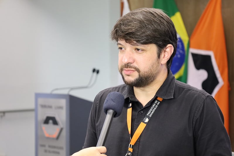 Homem com cabelo curto e barba, falando ao microfone em um evento público na Prefeitura de Divinópolis, com bandeiras ao fundo.