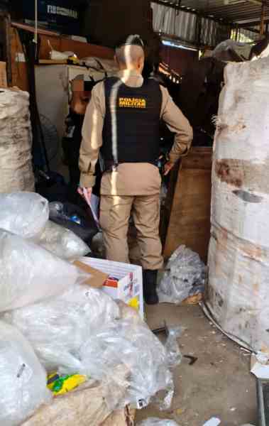 Policial da Polícia Militar examinando área desordenada durante operação contra comércio ilegal de fios de cobre.