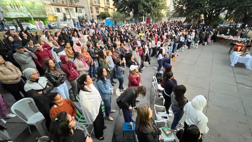 A large crowd of people gathered outdoors, many standing and some seated, during an event. The scene includes a backdrop of buildings and trees, with a few tables set up in the foreground.