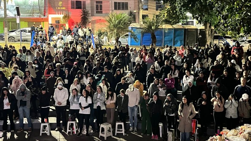 A large crowd of people gathered outdoors at night, some seated on stools, with various groups wearing winter clothing, under trees and streetlights.