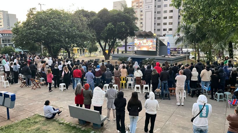 A large crowd gathered outdoors, facing a screen set up on a stage in a park. People are standing, some sitting on plastic chairs, with various clothing styles, including jackets and hoodies, indicating a cool environment.