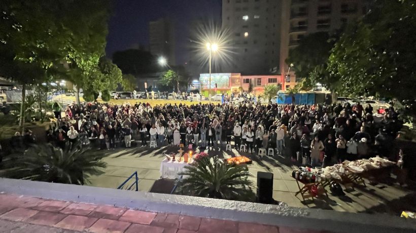 A large crowd of people gathered outdoors at night, with some attendees raising their arms. In the foreground, there is a table with offerings and decorations, surrounded by palm trees.