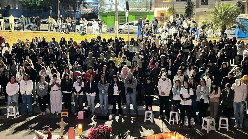 A large crowd of people gathered in an outdoor setting at night, many wearing warm clothing, with some individuals standing on stools. Candles and floral arrangements are in the foreground, hinting at a commemorative event.