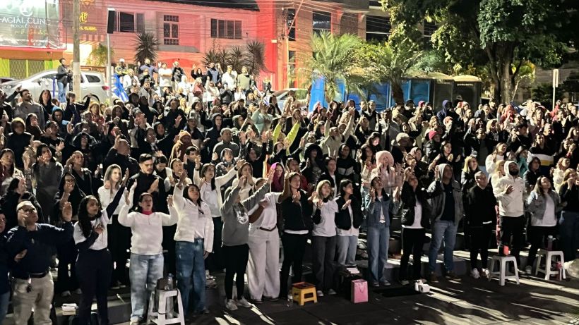 A large crowd of people gathered outdoors at night, with many individuals raising their hands in a communal display, illuminated by colorful lights in the background.