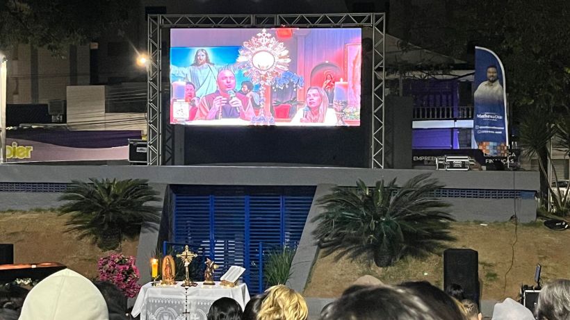 A large outdoor screen displaying a religious event with participants engaged in prayer, surrounded by decorations and plants.