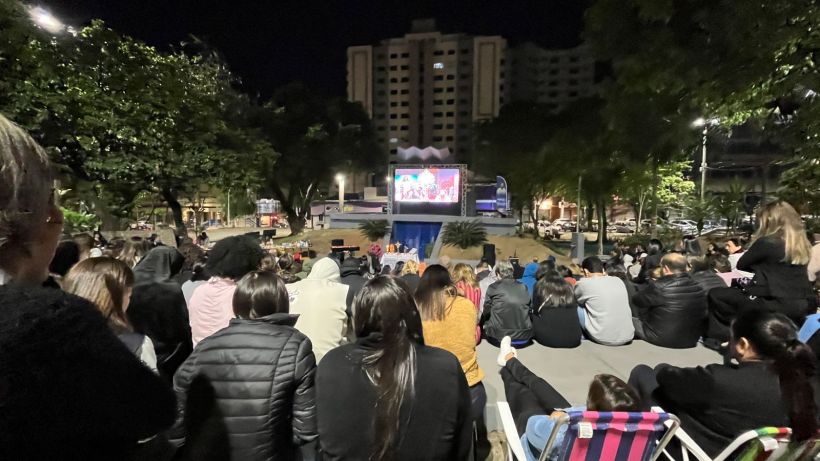 A crowd of people sitting on steps in a park, watching a large screen displaying a movie or event at night.