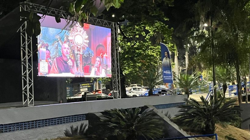 An outdoor screen displaying a religious event, featuring a speaker and audience in a lively park setting, surrounded by plants and illuminated by ambient lighting.
