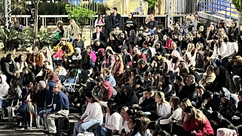 A large crowd of people gathered outdoors, sitting in bleachers and on the ground, some wearing jackets and scarves, engaged in an event at night.