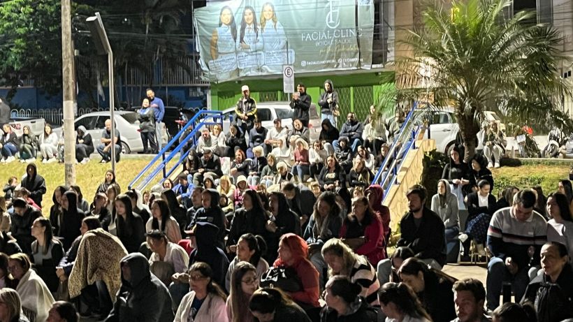 A large crowd of people gathered outdoors at night, sitting on stairs and the ground, some wearing hooded clothing. There is a banner in the background and a few cars parked nearby.