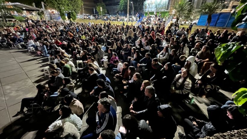 A large crowd of people sitting outdoors at night, listening to an event or performance. The audience appears engaged and attentive.