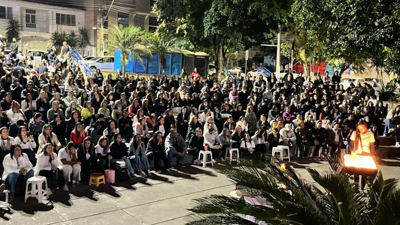 A large crowd of people seated outdoors, gathered together for an event at night, with palm plants in the foreground.