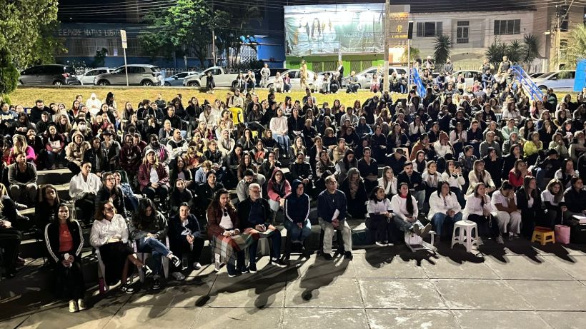 A large crowd of people gathered in an open space, sitting on steps and various seats, during nighttime. The scene captures a diverse audience, including individuals of different ages and backgrounds, focused in one direction, likely watching a performance or event.