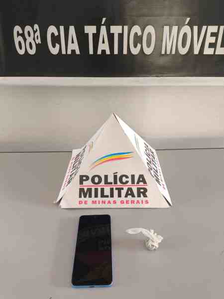 Um triângulo de papel com a marca da Polícia Militar de Minas Gerais e um celular em uma mesa, com um fundo que mostra texto em letras brancas.